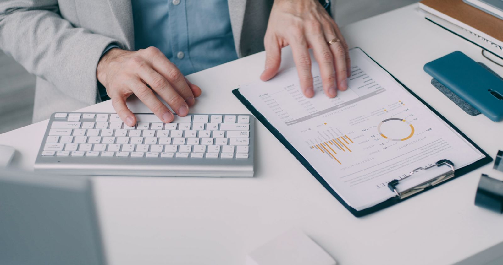 Finance professional at a keyboard reviewing a printed budget-versus-actual summary on a clipboard