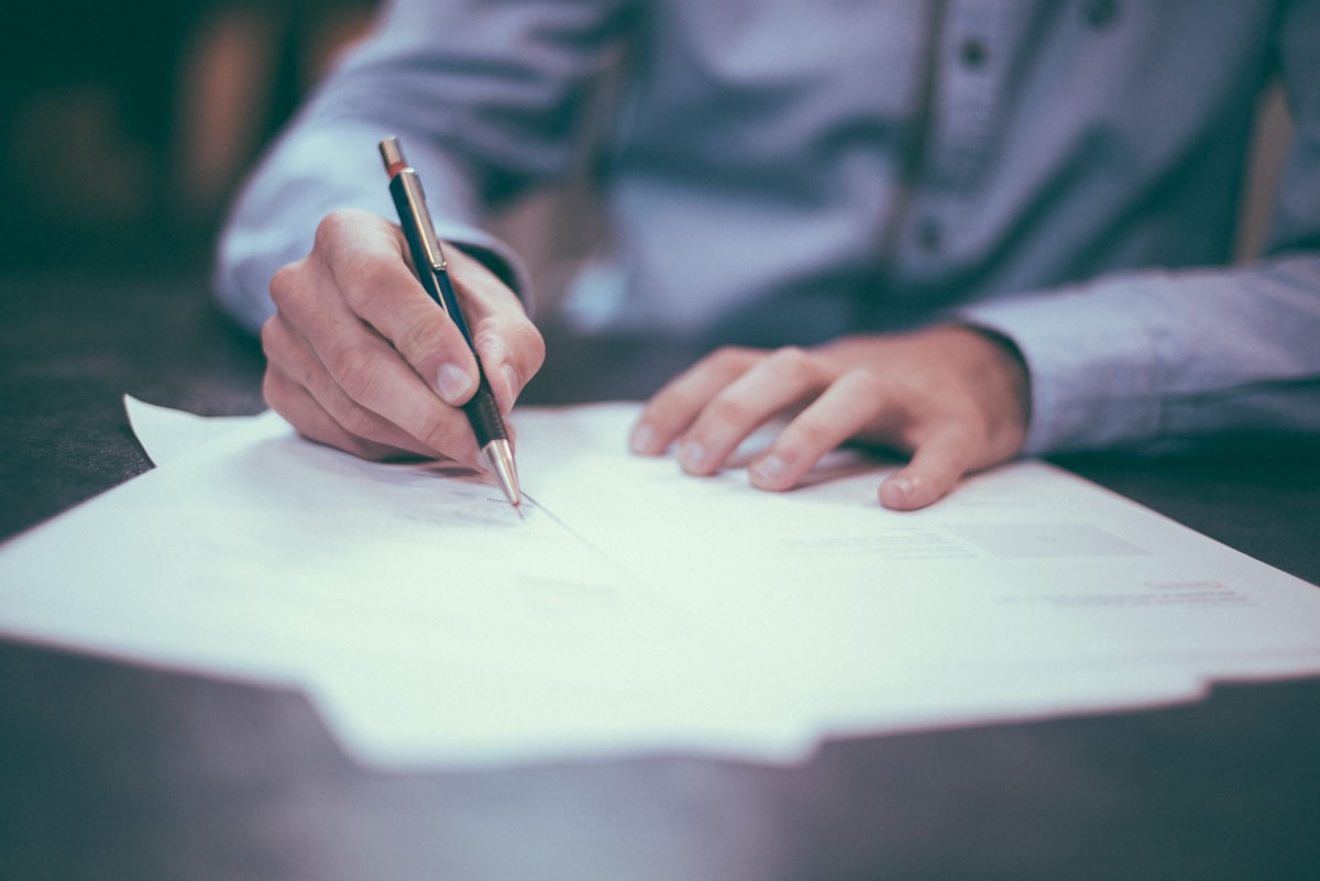 A person in a light blue shirt signing invoice paperwork with a pen at a desk, representing accounts receivable and collections work for a startup