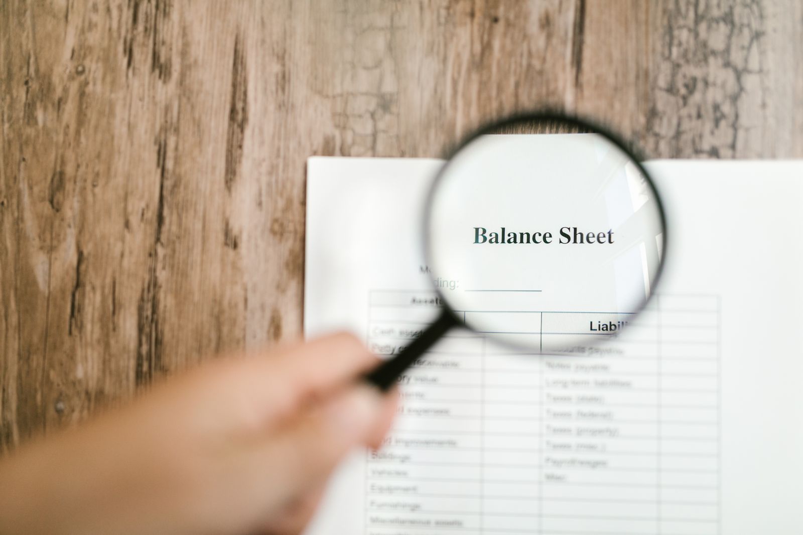Hand holding a magnifying glass over a printed financial statement on a wooden desk while reviewing the figures line by line