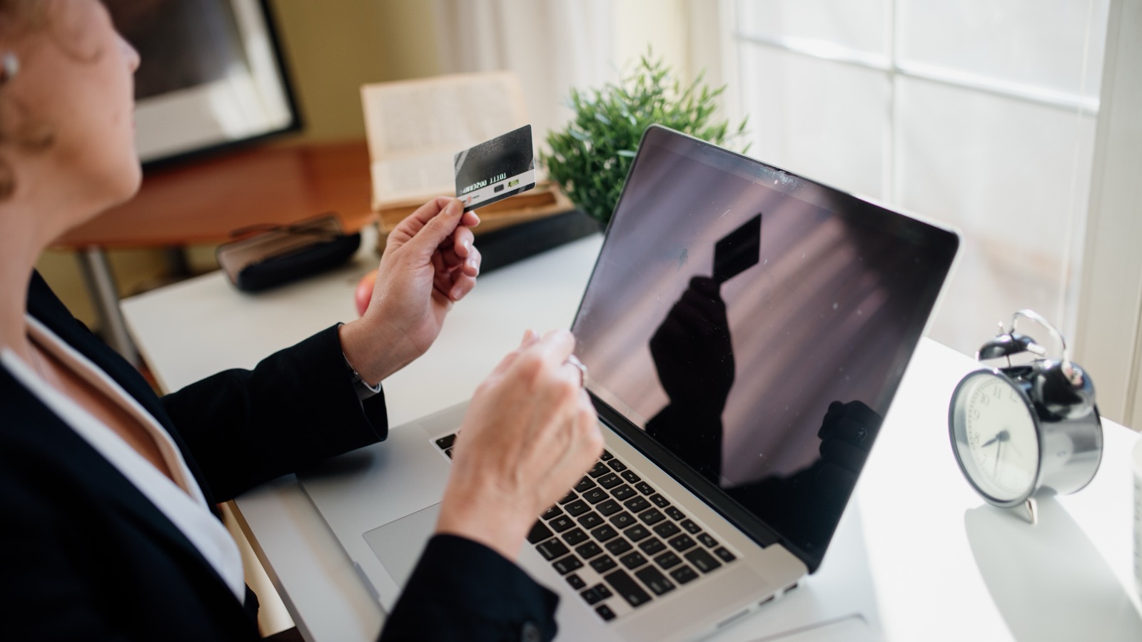 A person holding a payment card beside an open laptop at a bright home office desk