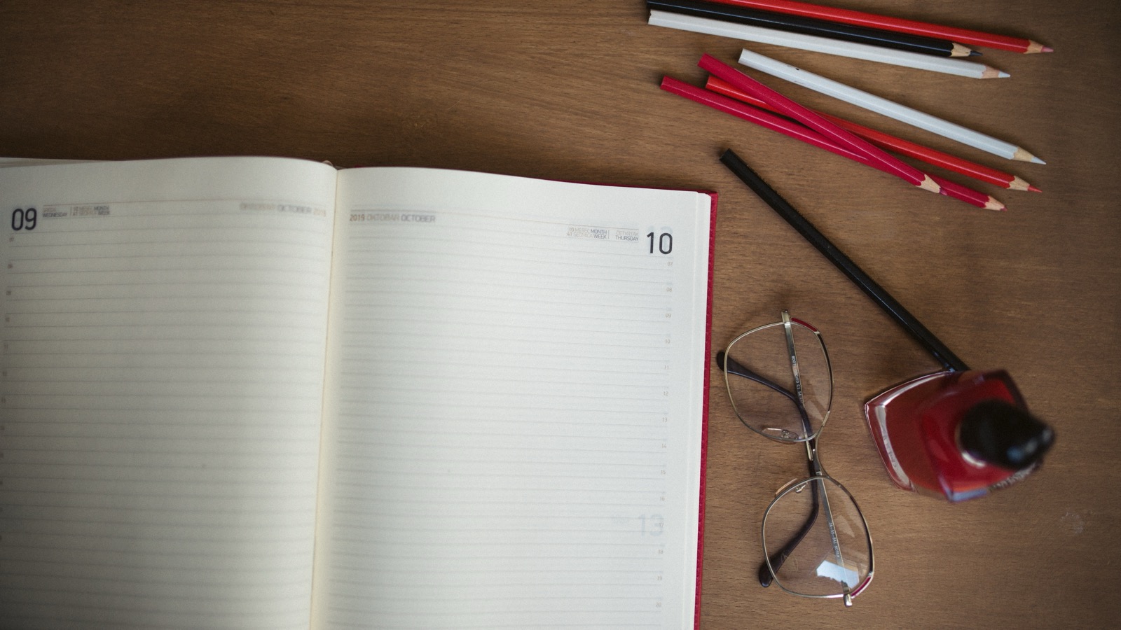 An open dated planner beside red and white pencils and reading glasses on a wooden desk